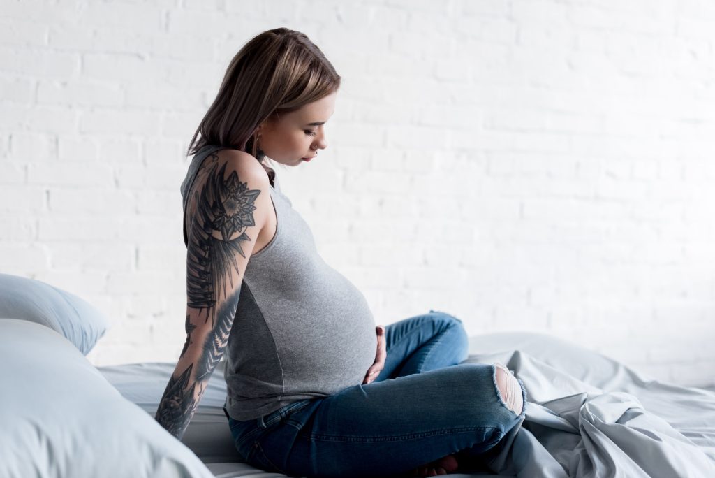 side view of pregnant tattooed woman sitting on bed at home