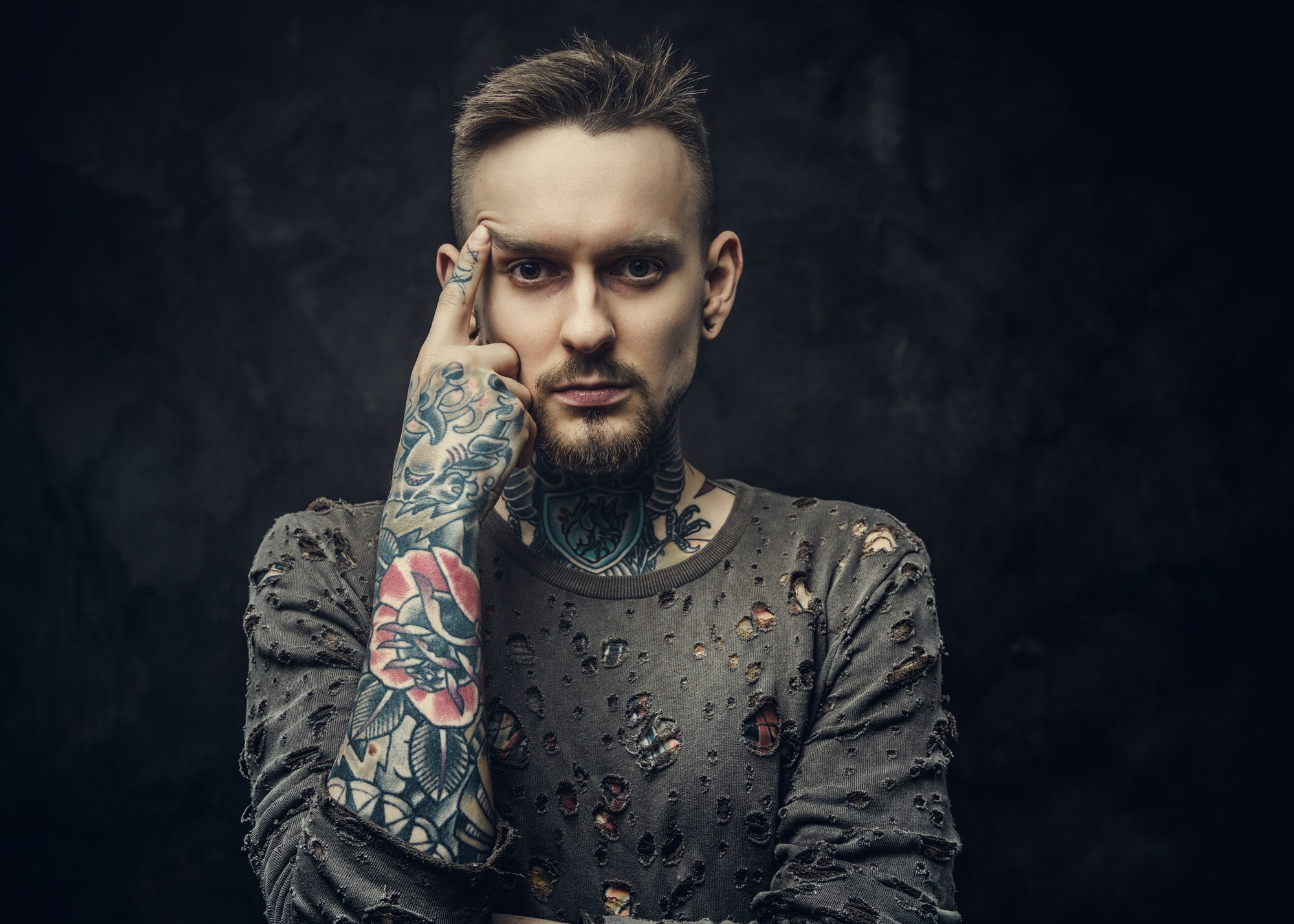 Tattooed male model posing in a dark grey background wearing ripped long-sleeved jumper